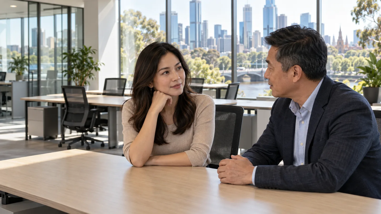 Two Vietnamese professionals talk at a desk in a sunlit Australian office with empty workstations behind them.