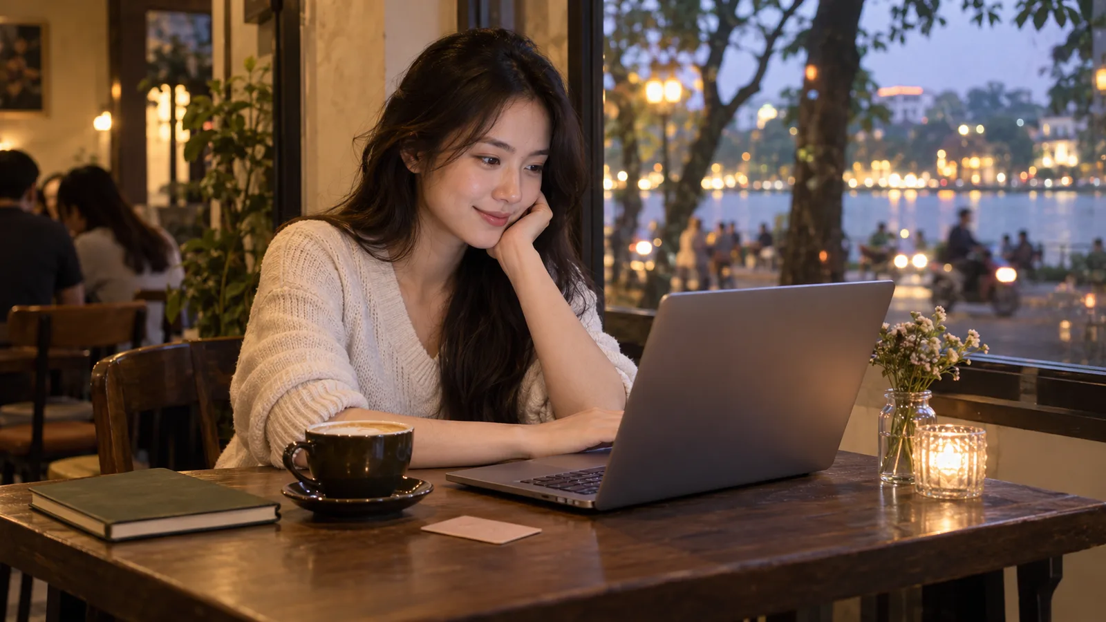 A young Vietnamese woman uses a laptop at a Hà Nội café, with a notebook and warm evening lights beside her.
